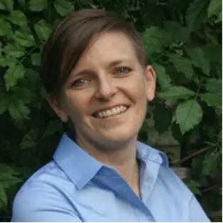 Person with short hair smiling, wearing a blue shirt. They are standing in front of lush green foliage.